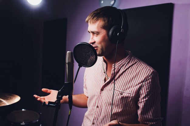 Music, show business, people and voice concept - male singer with headphones and microphone singing song at sound recording studio.