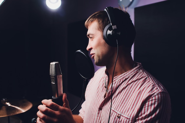 Music, show business, people and voice concept - male singer\
with headphones and microphone singing song at sound recording\
studio.