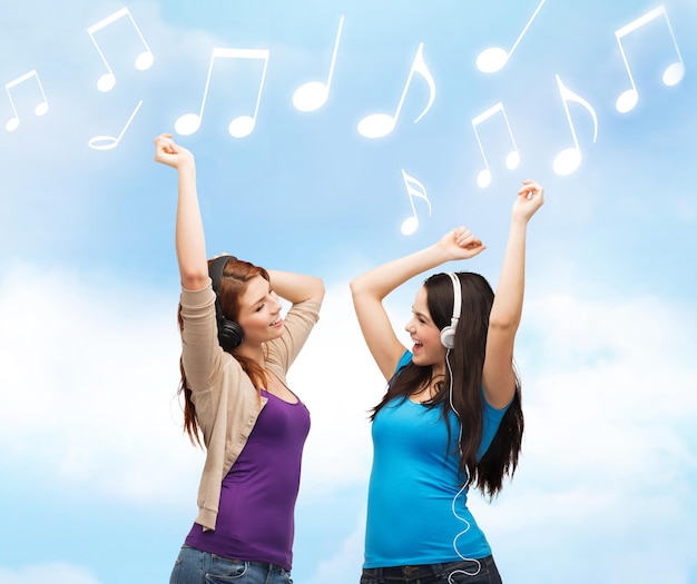 music and technology concept - two laughing teenagers with headphones dancing over blue sky background and music notes