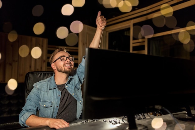 music technology gesture and people concept happy man at mixing console in sound recording studio showing thumbs up over lights