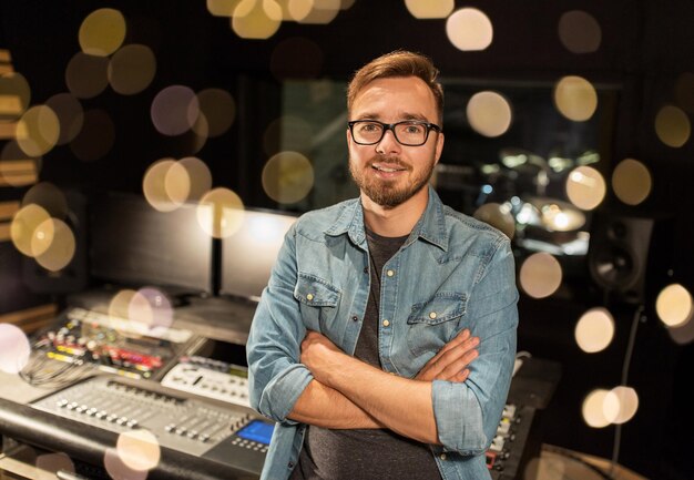 music technology people and equipment concept happy smiling man at mixing console in sound recording studio over festive lights