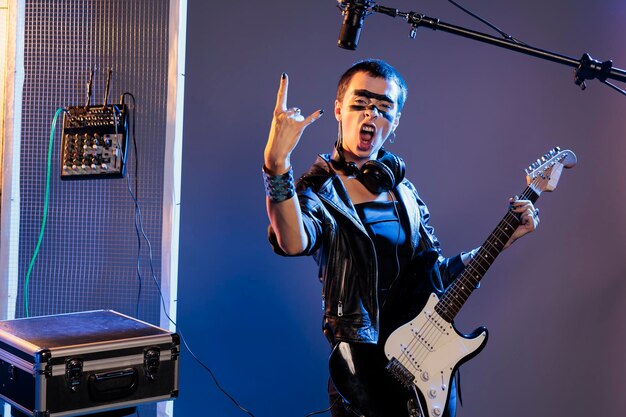 Musical artist doing rock sign and playing guitar, feeling ecstatic and doing solo concert in studio over background. female singer showing rock and roll symbol at live show performance