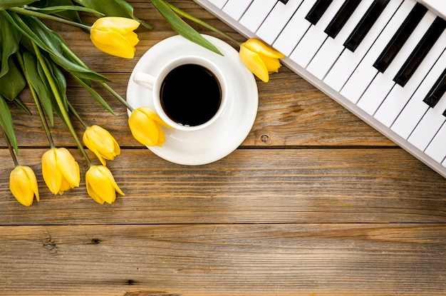 Musical background with white piano keys a cup of black coffee and a bouquet of yellow tulip flowers on a wooden surface top view copy space