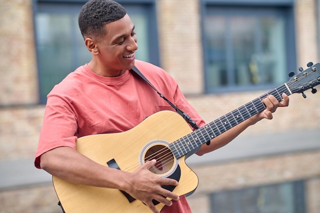 Musicista. giovane adulto sorridente dalla pelle scura in maglietta rossa che suona la chitarra in piedi sullo sfondo dell'edificio della città in una bella giornata