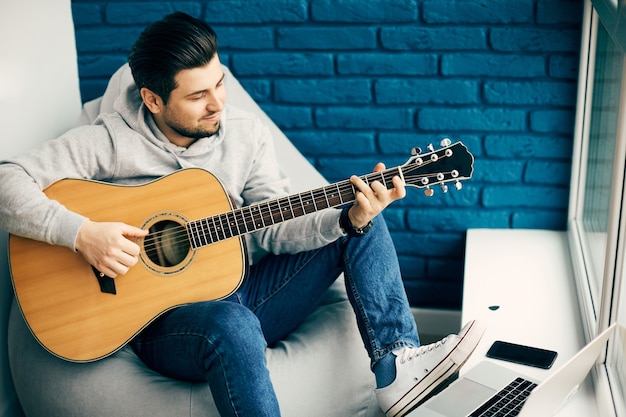 Musician playing on fresh new guitar at home with laptop, happy guitarist practicing with bue bricks