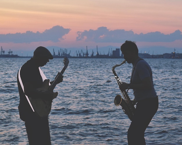 Photo musician playing musical instruments while standing against sea during sunset