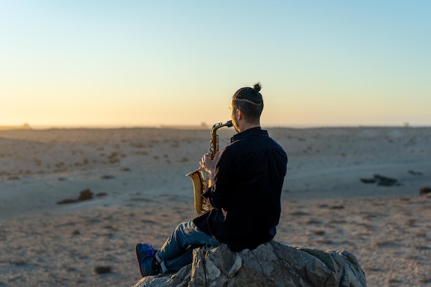 Музыкант сидит на скале и играет на саксофоне на закате в пустыне