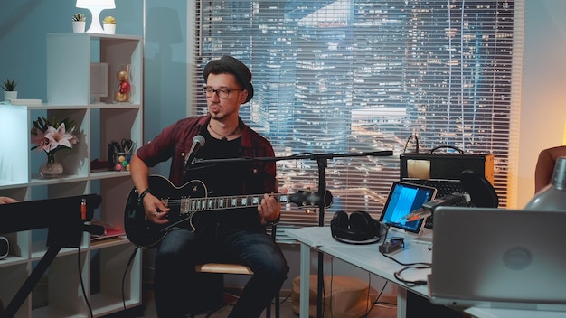 Musician trying sound recording equipment in home studio in the evening. He singing and playing guitar while his friend accompanying him.