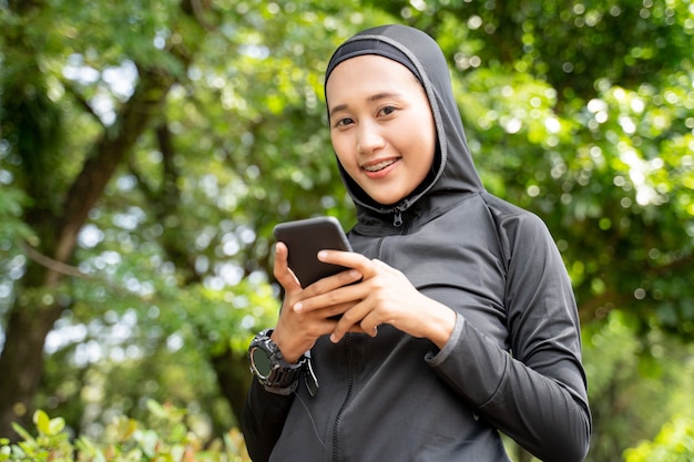 Азиатская мусульманская женщина улыбается, используя свой смартфон во время занятий спортом на открытом воздухе