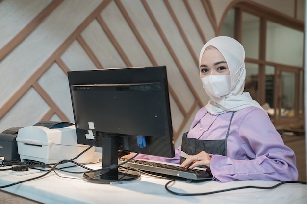 사무실에서 보호를 위해 의료 얼굴 마스크를 착용하는 동안 PC를 사용하여 작업하는 이슬람 아시아 여자