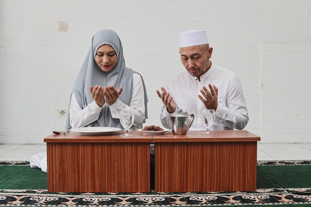 금식 iftar 저녁 식사를 함께 하기 전에 기도하는 이슬람 부부는 손을 들고 고개를 숙입니다