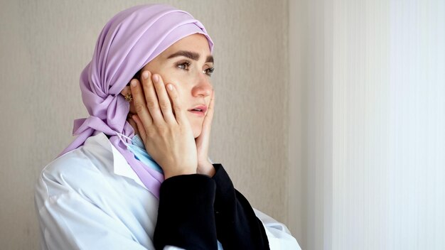 ピンクのヒジャブと作業服を着て窓の外を眺めるイスラム教徒の医師が頭を抱える 女性は仕事で疲れて動揺した表情をしている