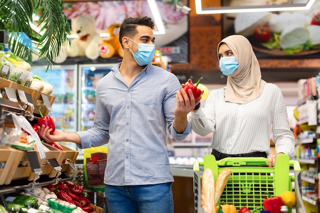 Coppia di famiglia musulmana che compra cibo scegliendo le verdure durante la spesa al supermercato, indossando maschere protettive. coniugi islamici che fanno la spesa nel fine settimana