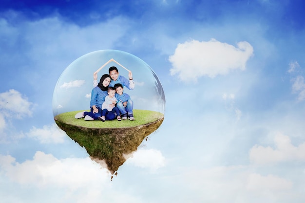 Muslim family holding a house roof symbol from cardboard over their heads while sitting on floating island with bubble protection in the blue sky