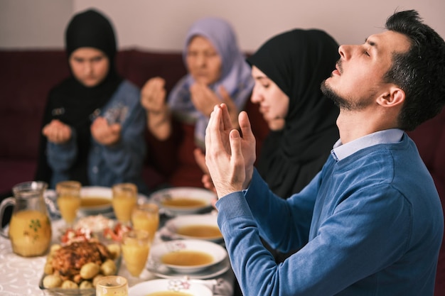 Muslim family making iftar dua to break fasting during Ramadan dining table at home together praying with hands up