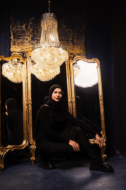 Muslim girl in black hijab near mirrors with her reflections Fashion muslim model near big expensive chandelier Islamic religion