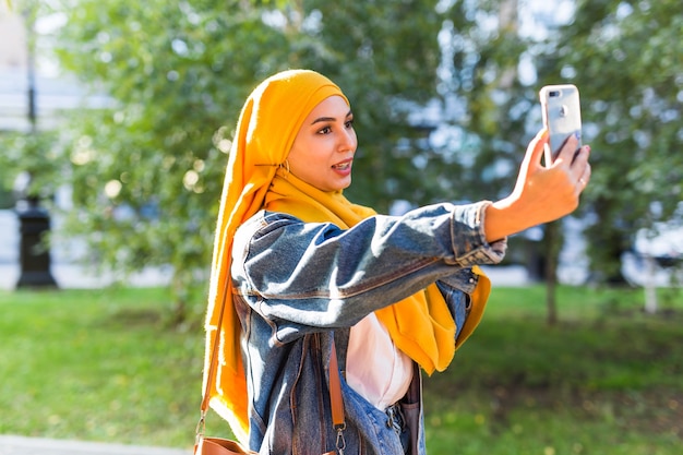 ヒジャーブのイスラム教徒の少女は、街の通りに立っている電話で自分撮りをします