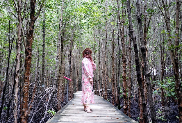 Muslim girl smiling to camera enjoy vacation at tropical sea.
teenager girl wearing sun straw hat and white dress standing on
wooden bridge with mangrove forest green plant background.