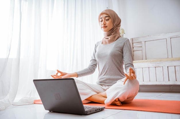 Muslim hijab woman watching home work out tutorial from laptop on a mattress in a room happily