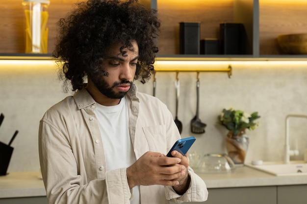 Muslim man watches photos in smartphone standing in kitchen