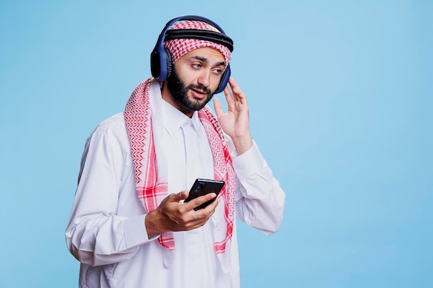 전통적인 옷을 입은 무슬림 남자가 무선 헤드폰으로 음악을 듣고 스마트폰을 사용하는 아랍인 이슬람 헤드 스카프를 입고 헤드폰으로 소리를 즐기고 휴대폰을 들고 있습니다.