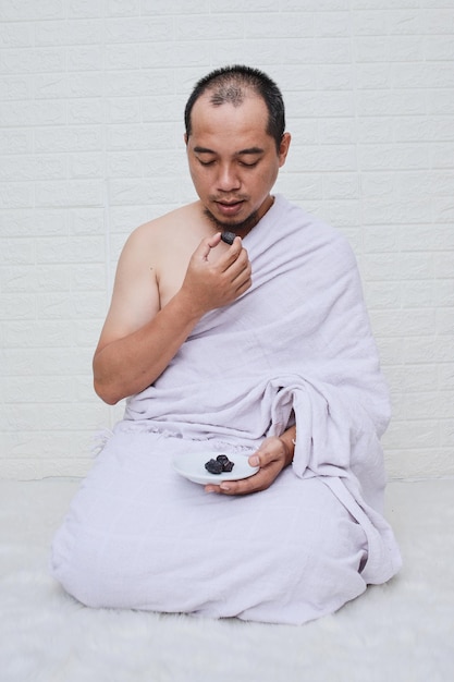 Muslim man wearing white ihram clothes, holding a bowl of dates fruit over white background