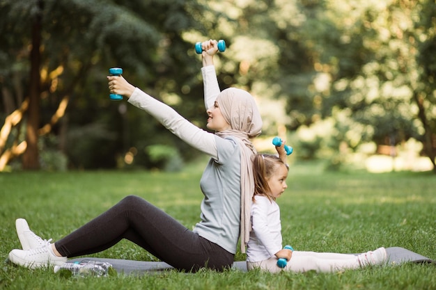 Muslim mother and her daughter sitting on mat and lifting dumbbells on fresh air at summer day