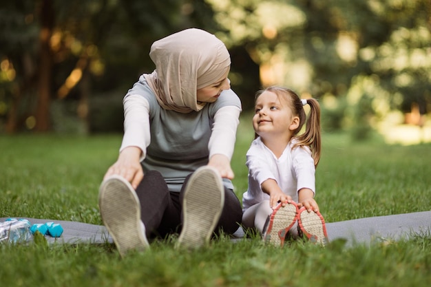 Muslim mother and her little daughter doing stretching exercises on yoga mat outdoors in the park