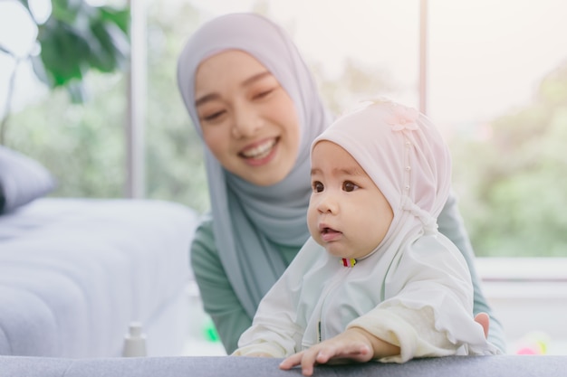 이슬람 어머니는 그녀의 아기가 행복한 미소 짓는 유아 가정 보살핌과 함께 귀엽고 사랑스러운 어린 시절을 실내에서 바라보고 있습니다.