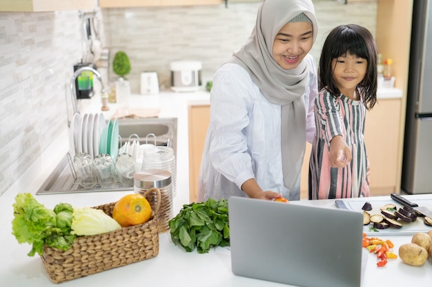 Muslim mother looking at recipe from laptop and cooking with her daughter. having fun woman with hijab and kid preparing dinner together