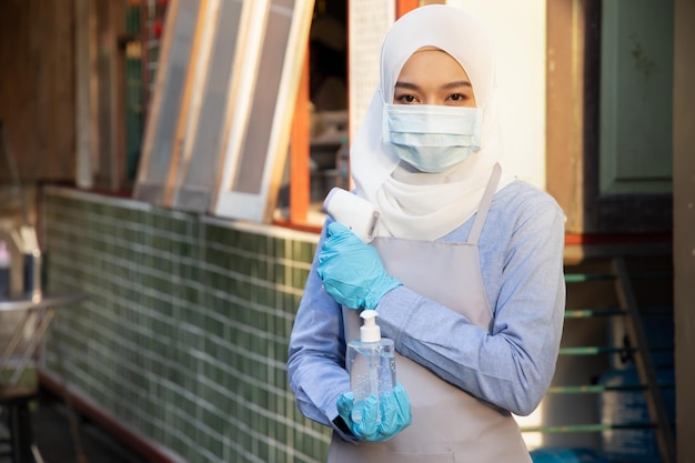 Foto impiegata musulmana o negoziante che tiene disinfettante per le mani con dispenser di gel alcolico e pistola termica per scanner per il controllo della temperatura nuovo concetto di pratica commerciale normale