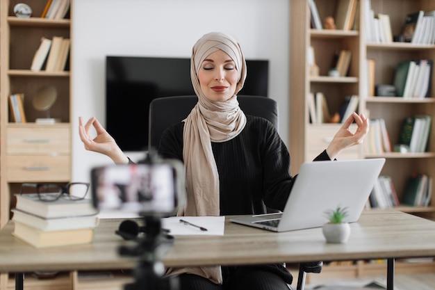 Donna musulmana libera professionista seduta al tavolo con gli occhi chiusi che registra sul blog come alleviare lo stress