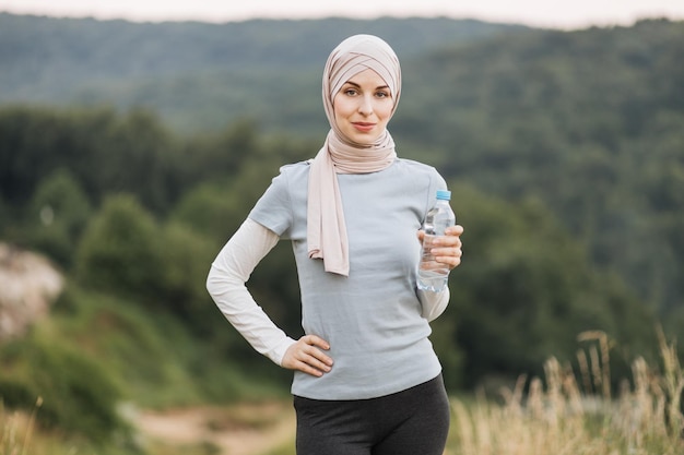 Muslim woman in hijab and sport clothes standing at green park and holding bottle of water