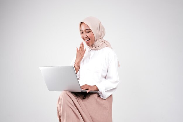 Photo a muslim woman is sitting on a chair and is using a laptop.