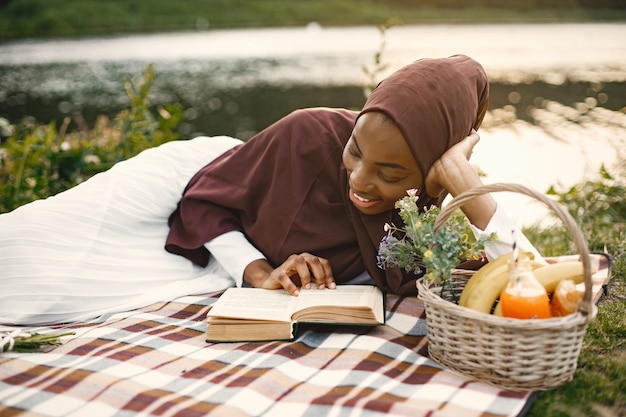 A muslim woman lay on the plaid picnic blanket near the river and reading a book