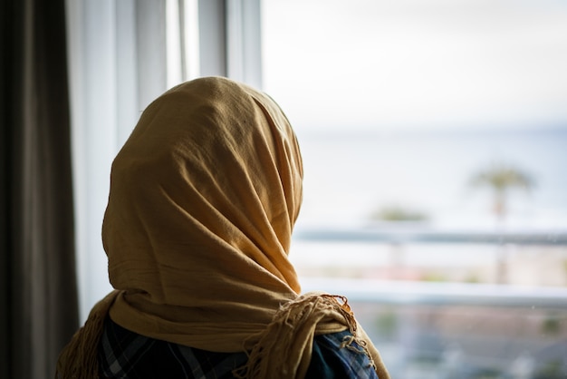 Muslim woman looking from hotel apartment window at the sea