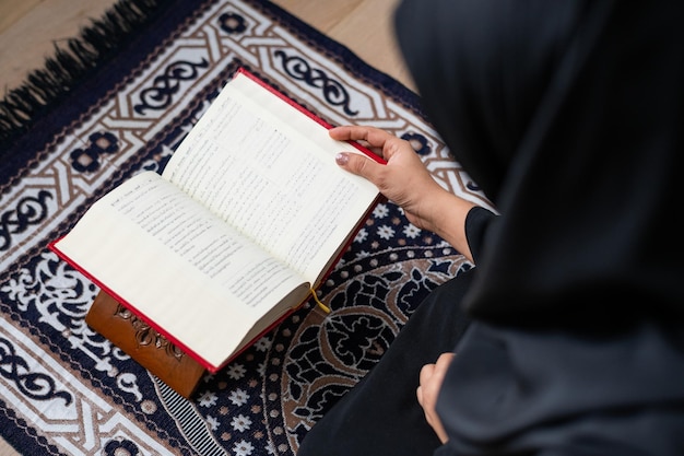 Muslim woman reading a book on a patterned rug The book is open and the person is wearing a black garment The setting is calm and focused on reading Muslim woman reading the Quran
