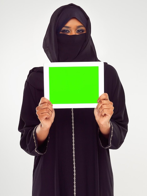 イスラム教徒の女性のタブレットと灰色のスタジオの背景に対する広告またはモックアップをマーケティングするための緑色の画面緑色のクロマキー画面またはディスプレイでタッチスクリーンを保持しているヒジャーブの女性の肖像画