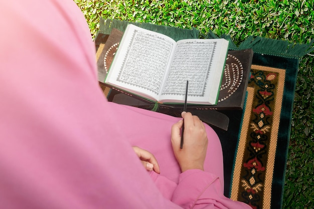 Muslim woman in a veil sitting on the prayer rug and reading the Quran at outdoor