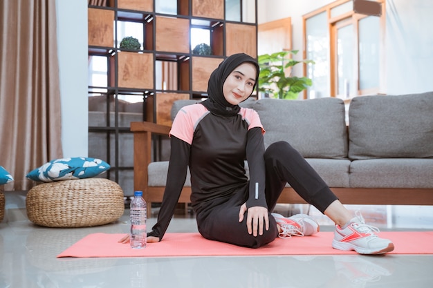 A Muslim woman wearing a hijab gym outfit sits casually by a water bottle on the floor at home