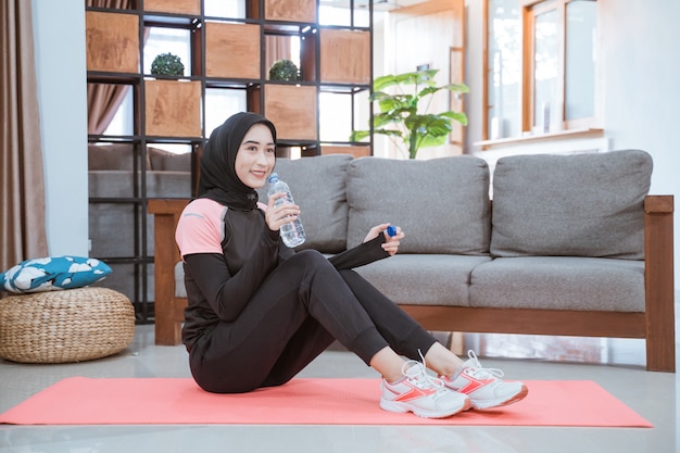 A Muslim woman wearing a hijab gym outfit sits on the floor holding a drinking water bottle in the living room