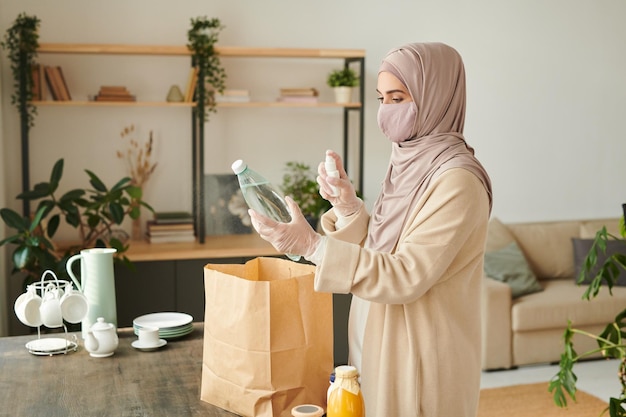 Donna musulmana che indossa una maschera protettiva hijab sul viso e guanti che disinfettano le merci a casa dopo essere andate