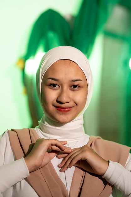 A Muslim woman in a white headscarf and white clothes poses with her hands without the slightest makeup on her face and smiles during a photo shoot