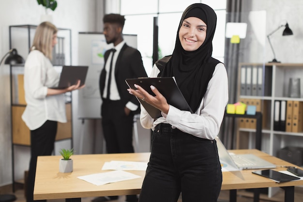Muslim woman with clipboard smiling on camera at boardroom