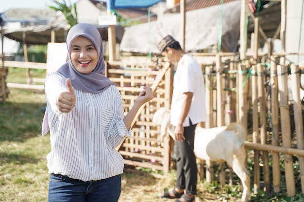 염소 농장에 서있는 동안 엄지 손가락을 보여주는 스카프와 무슬림 여성. eid adha 개념