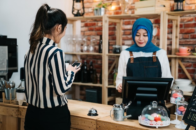 イスラム教徒の女性労働者が喫茶店で顧客の注文を受けます。カフェバーで若いビジネスオフィスの女性の朝のルーチンは、スマートフォンの支払いカウンターを使用して朝食を購入します。笑顔のヘッドスカーフでイスラムの女の子のウェイトレス