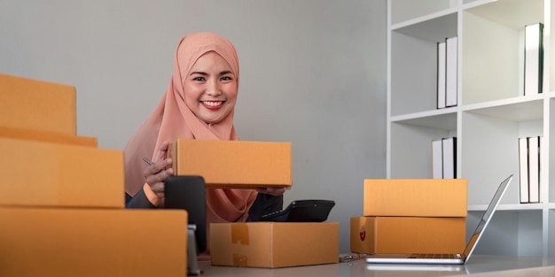 Muslim women selling online at home with box Selling online with box to accept order from customer SME business idea Parcel delivery muslim woman working smartphone and laptop at home