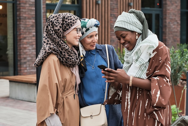 Muslim women talking to their friend while she reading a message on the phone and smiling during their meeting outdoors