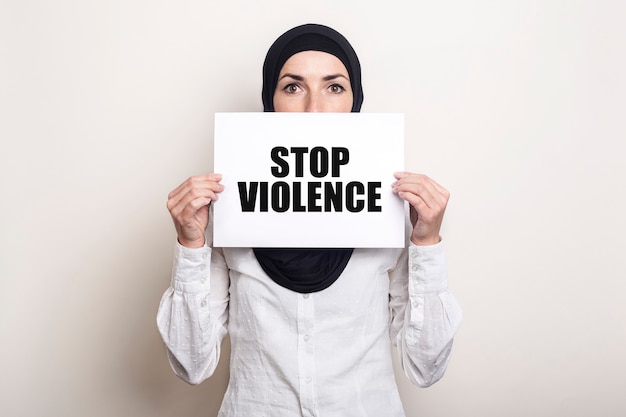 Muslim young woman in hijab covers her face with a banner with the text STOP VIOLENCE.