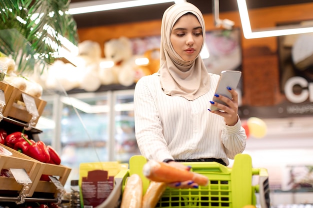 食料品店で携帯電話を使用して買い物をするイスラム教徒の若い女性